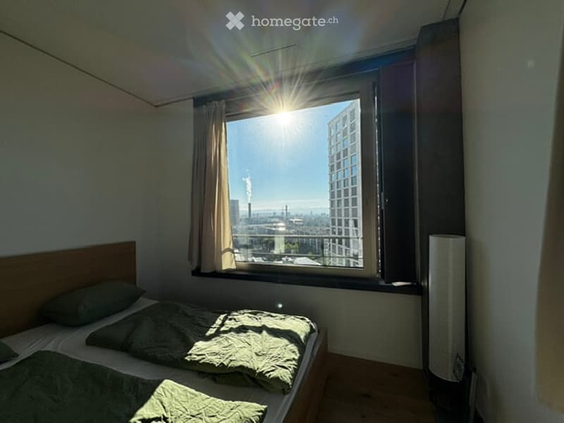 Bright bedroom with a single bed, pillows, and a large window offering a city view. The room features a radiator and curtains.