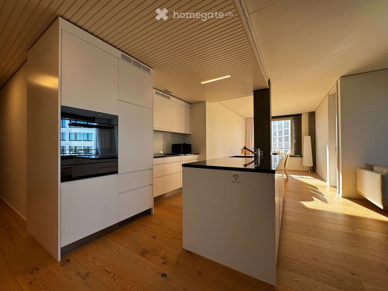 Modern kitchen with white cabinets, a central island, and a sleek black countertop. The space features wooden flooring, built-in storage, and a small seating area with a sofa and stools.