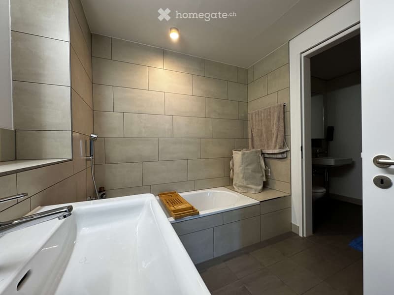 Modern bathroom with a large bathtub, tiled walls, a mirror, a towel, and a wooden tray on the edge of the tub. A beige bag is placed next to the towel. The bathroom has a sleek design with a visible light fixture on the ceiling.