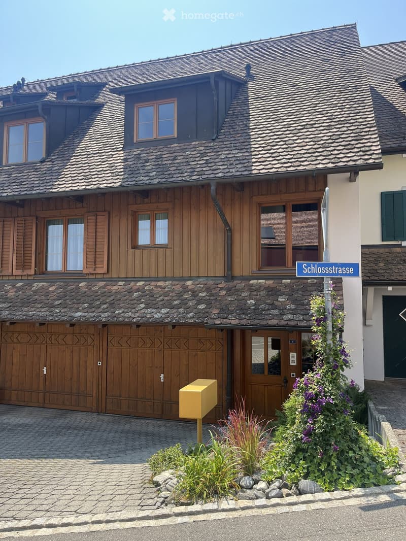 Two-story house with a steep, multi-pitched roof, wooden facade, stone lower section, and large wooden garage doors. Features multiple windows with shutters and a small garden with plants and flowers in front.
