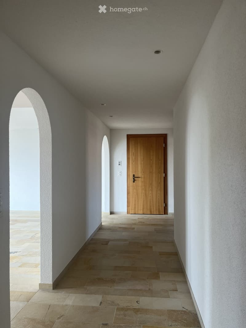 A modern hallway with beige tiled flooring, white textured walls, and a wooden door at the end. The ceiling has recessed lighting and a slightly arched design.