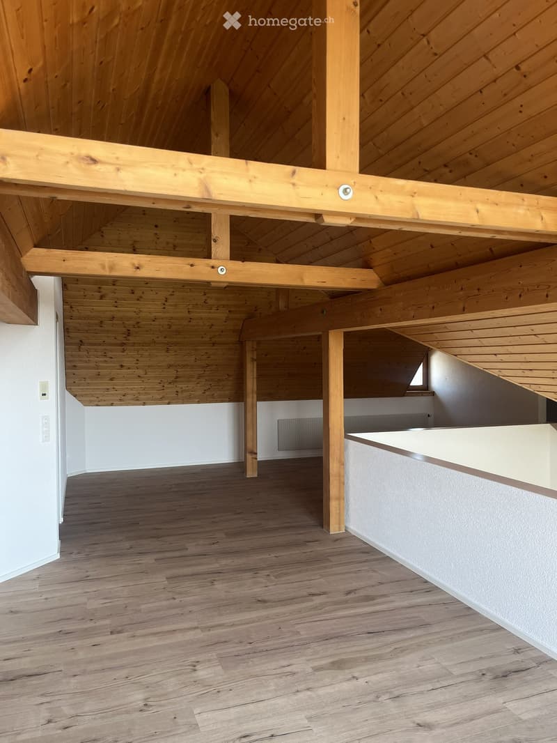 Empty room with wooden floor and exposed wooden beams on the ceiling. White walls and a small window visible in the background.