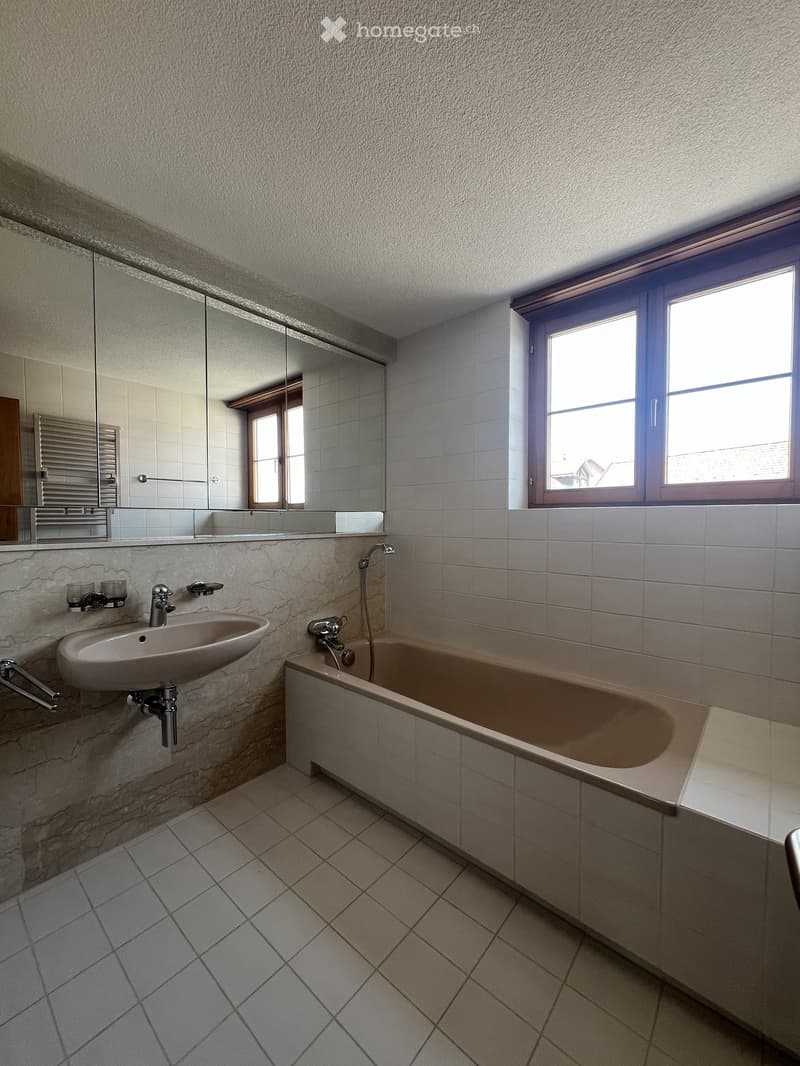 Modern bathroom with a bathtub, sink, mirror, and two windows letting in natural light. The walls are tiled, and the floor has a diamond-patterned tile design.