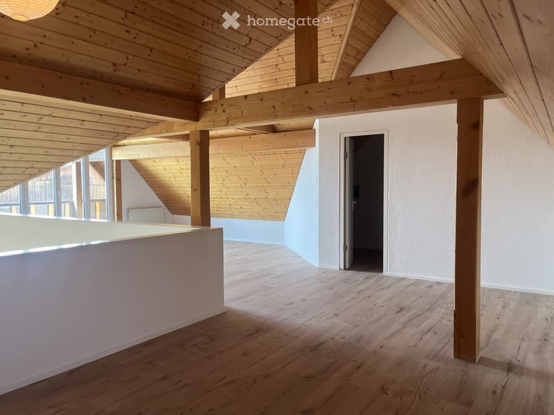 Empty room with wooden flooring, exposed wooden beams, and a white partition wall. The room has large windows and a doorway leading to another space.