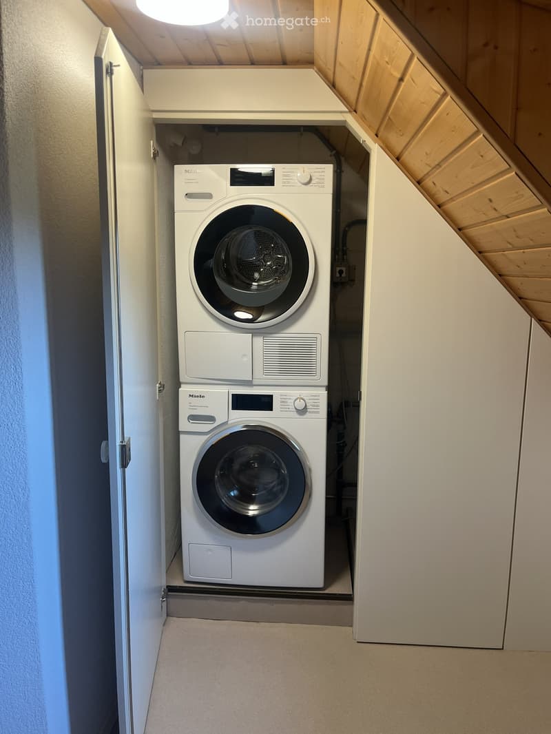 Compact laundry area with a stacked washing machine and dryer unit, white cabinets, and wooden slanted ceiling.