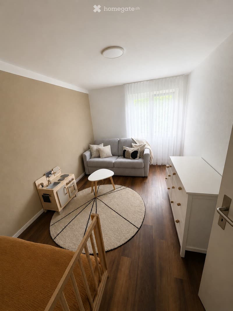 A small, cozy living space with a grey sofa, decorative pillows, a round white stool, a toy kitchen, a white cabinet, and a round patterned rug on a wooden floor.