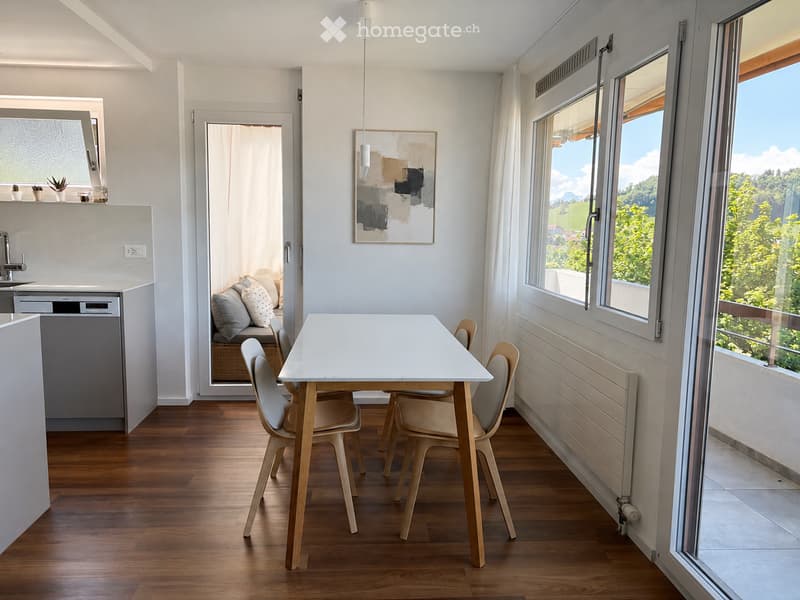 Bright, modern open-plan living and dining area with wooden flooring, large windows offering a scenic view, a white dining table with four chairs, a small kitchen area with a dishwasher and sink, and a cozy seating area with a sofa and pillows.