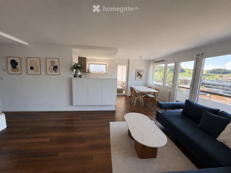 Modern living and dining area with hardwood floors, large windows offering a scenic view, a blue sofa, a white coffee table, a dining table with chairs, and a white kitchen cabinet.