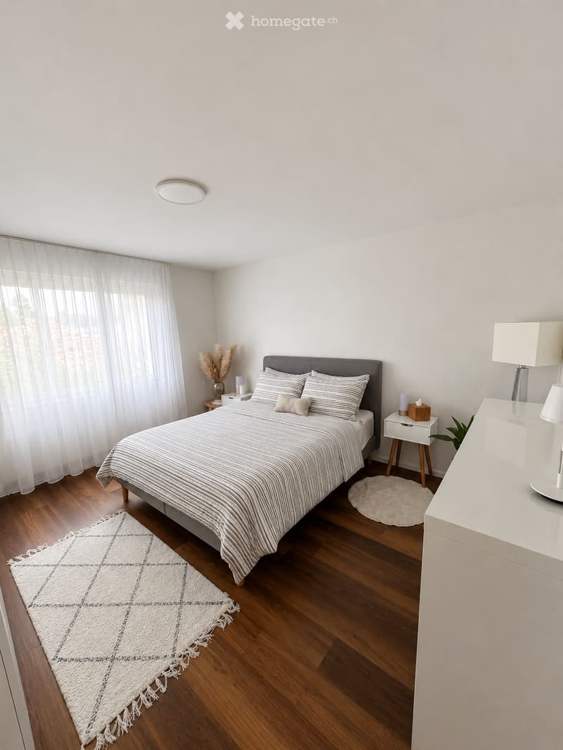 Modern bedroom with a large bed, nightstand, lamp, and decorative rug. The room features wooden flooring and large windows with sheer curtains.