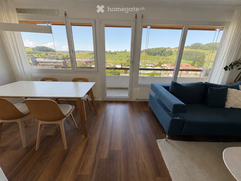 Modern living room with wooden flooring, large windows offering scenic views, a blue sofa, a white dining table, and chairs.