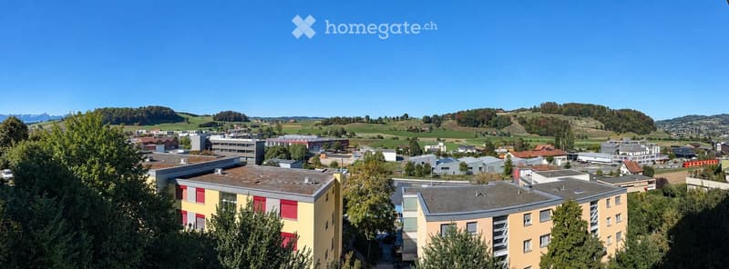 A panoramic view of a suburban area with multiple buildings, green fields, and distant mountains under a clear blue sky.