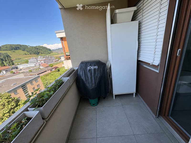 Balcony with tiled floor, large window, view of greenery and town, refrigerator, plant pots, and a covered barbecue.