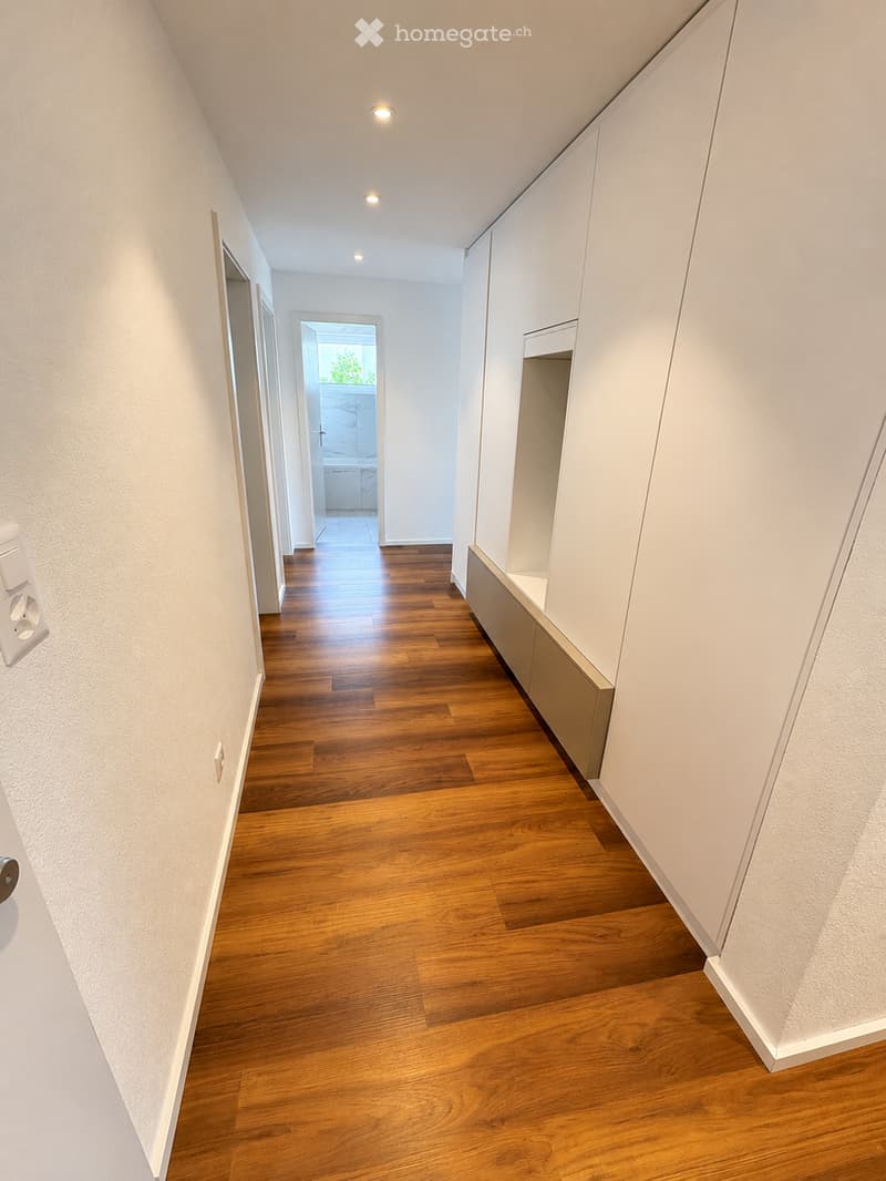 A modern hallway with wooden flooring, white walls, built-in storage cabinets, and recessed lighting. The hallway leads to an open area with a view of a green space.