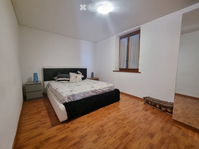 Bedroom with wooden flooring, white walls, large window with curtains, bed with headboard, nightstand, and decorative items.