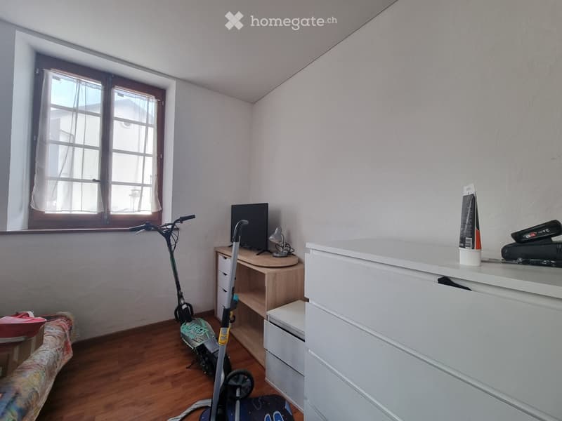 A small, furnished room with a wooden floor, a desk with a screen and lamp, a scooter, and a bed with a colorful blanket.