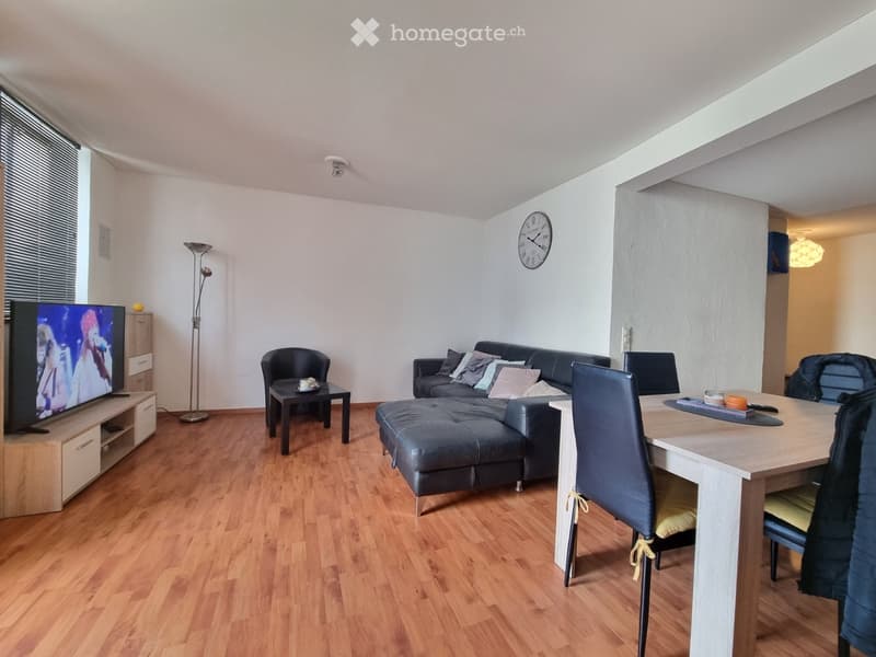Living room with hardwood flooring, a sectional sofa with an ottoman, a coffee table, a dining table with chairs, a television, a floor lamp, and a clock on the wall.