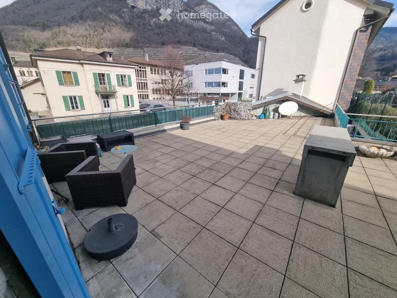 Balcony with tiled floor, outdoor furniture including chairs, a table, and a stool, overlooking a town with mountains in the background.
