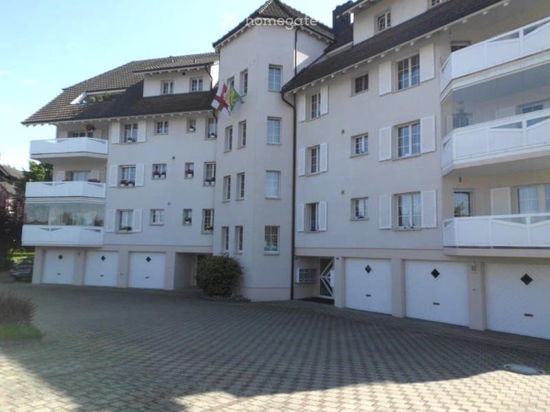 Multi-story residential building with white exterior, multiple balconies, garage, and parking area.