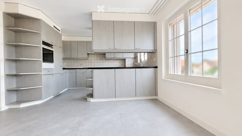 Modern, unfurnished kitchen with light grey cabinets, a central island, and large windows allowing natural light.