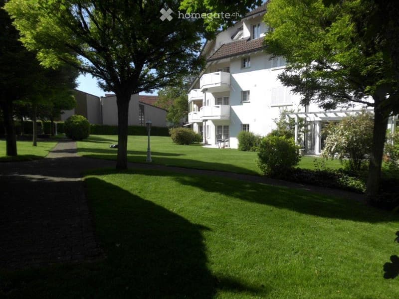 Well-maintained garden area with a large white building featuring balconies, surrounded by trees and shrubs.
