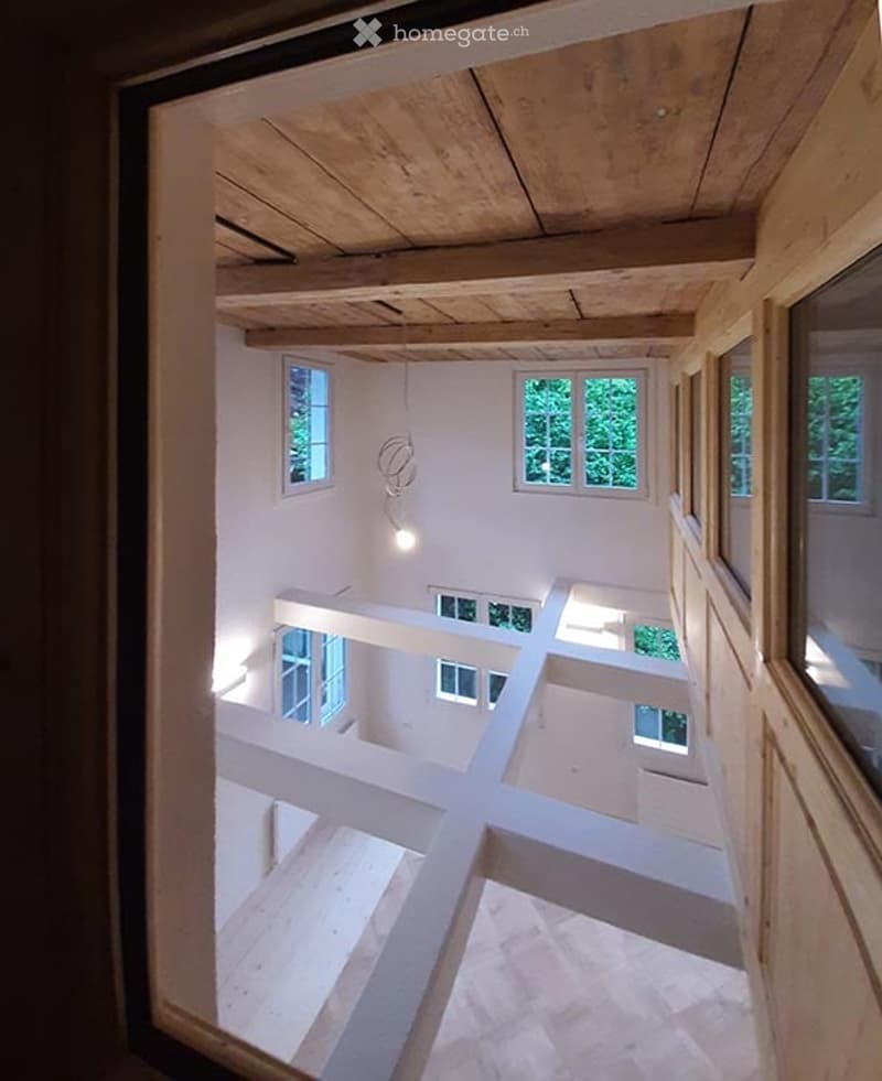 Open, airy interior space with exposed wooden beams and white structural supports. Multiple windows allow natural light to fill the room, and the floor appears to be made of light-colored wood or concrete.
