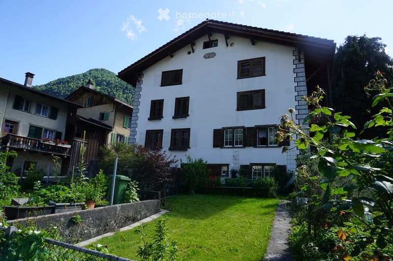 A two-story white house with brown shutters and a sloped roof, surrounded by a well-maintained garden and greenery, with a mountain view in the background.