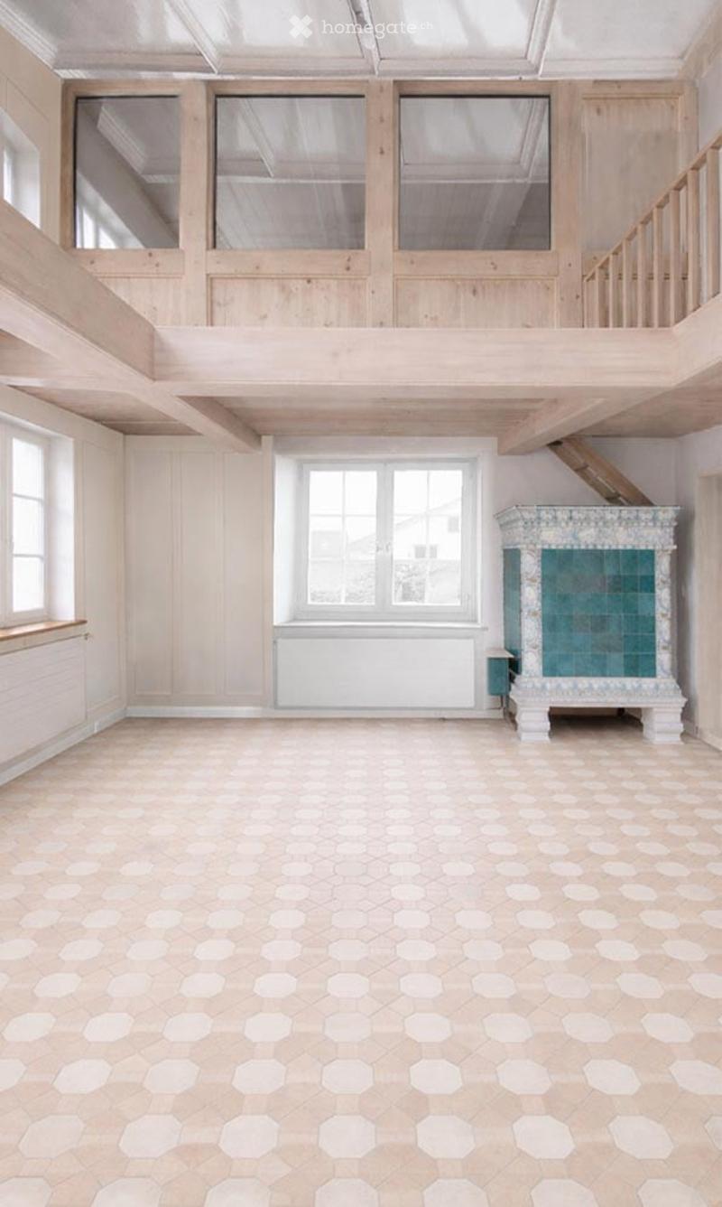 Empty room with exposed wooden beams, large windows, and a fireplace with a decorative tile surround.