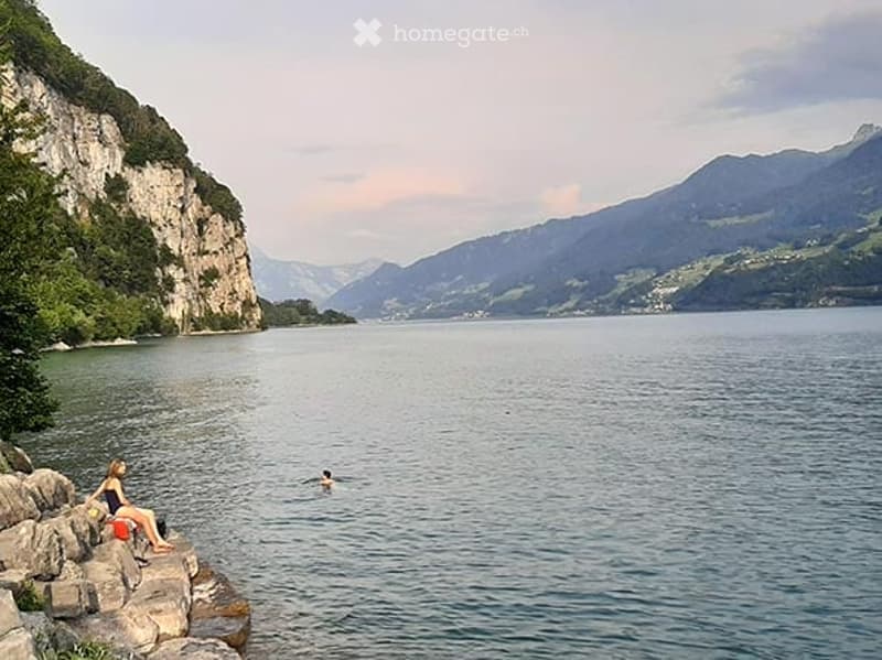 A serene lakeside scene with calm water, rocky shore, and distant mountains. Two people are present, one sitting on a rock and the other swimming in the lake.