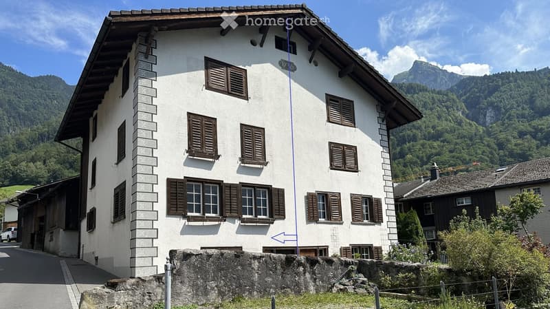 A two-story white house with brown shutters and a stone base, situated in a mountainous area with lush greenery and a clear blue sky.