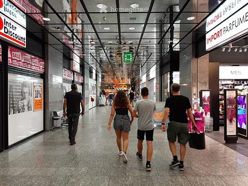 A busy shopping mall corridor with people walking, various store signs, and a modern ceiling with recessed lighting.