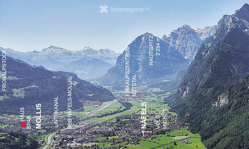 A panoramic view of a valley surrounded by mountains, with a town nestled in the valley floor and green fields interspersed with forested areas.