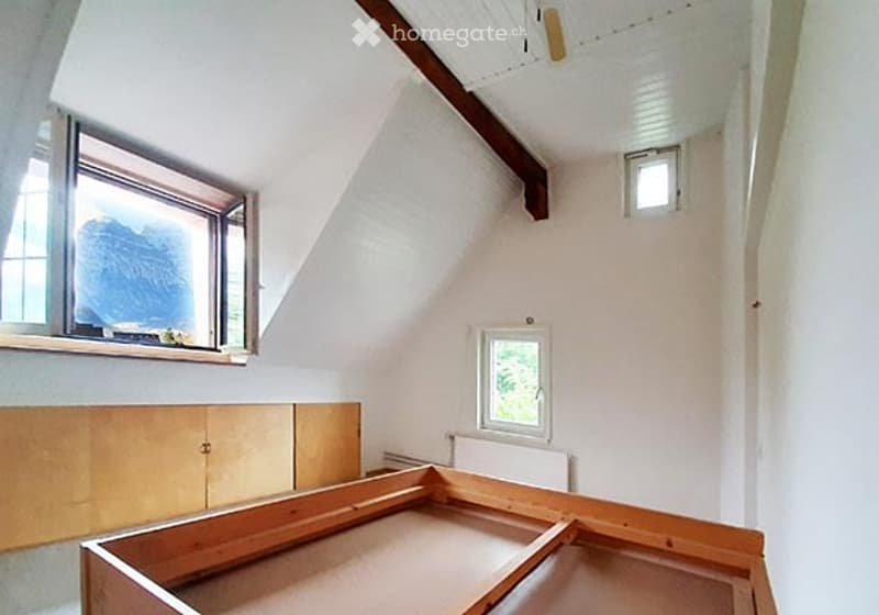 An attic room with exposed wooden beams, white walls, and large windows. The space is empty, showing the wooden floor and built-in wooden cabinetry.