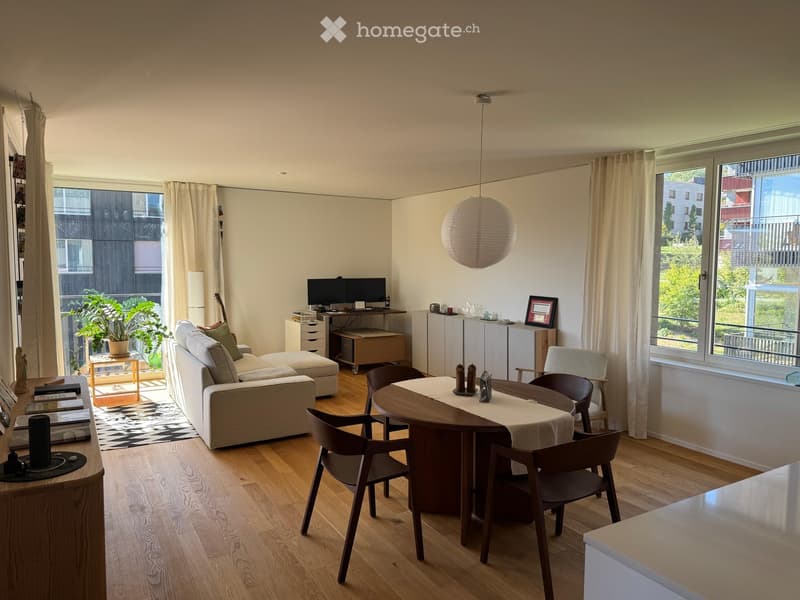 Bright living room with wooden flooring, large windows, a sofa, ottoman, coffee table, dining table with chairs, laptop on a desk, and a potted plant near the window.