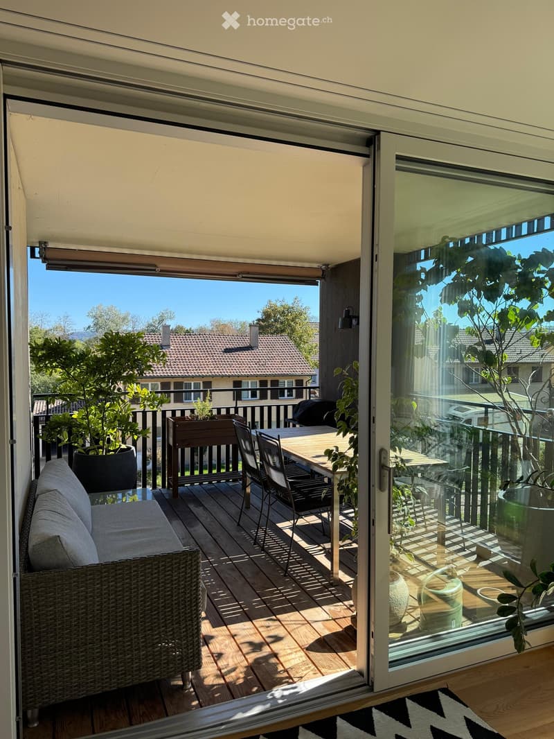 Balcony with wooden decking, sofa, dining table, chairs, and potted plants, offering a scenic view of the surrounding area.