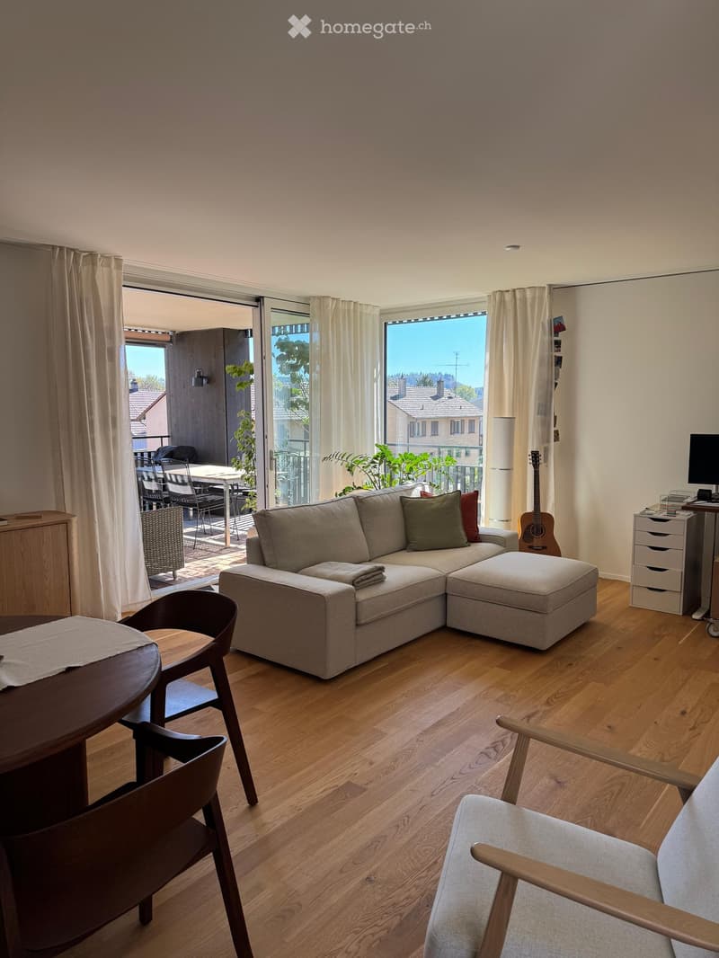 Modern living room with wooden flooring, large windows, and a balcony. Features a sectional sofa, wooden dining table, chairs, and a guitar.