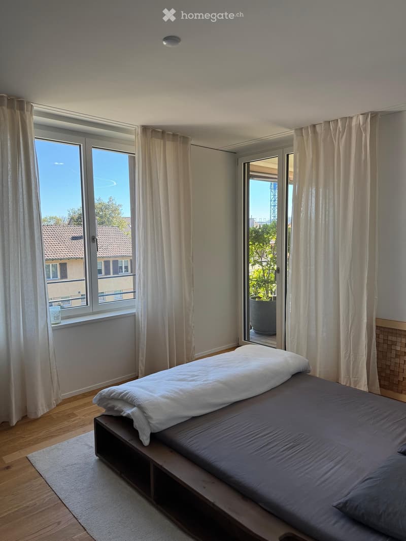 Modern bedroom with a wooden bed, grey bedding, large windows with curtains, and a view of the city.