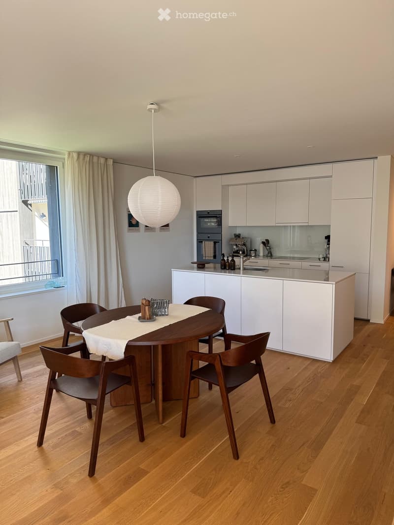 Modern kitchen with white cabinets, wooden dining table, four chairs, and a hanging lamp. The kitchen features an oven, coffee maker, and various small appliances on the counter.