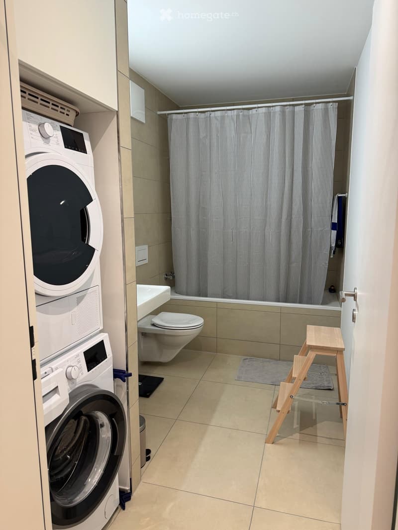 Modern bathroom with a washing machine, toilet, sink, shower curtain, and a small wooden stool.