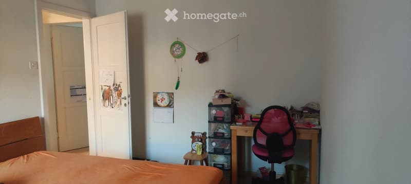 Cozy bedroom with a wooden bed, a small desk with a red office chair, a storage unit, and a decorative toy on a stool. The room features a calendar, a colorful mobile, and an open door leading to another room.
