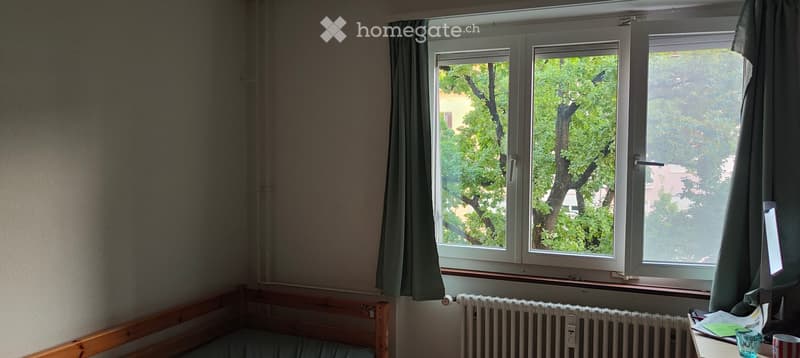 A small, furnished bedroom with a wooden bed frame, a bedside table with a lamp, a radiator, and a window with curtains overlooking a leafy tree.
