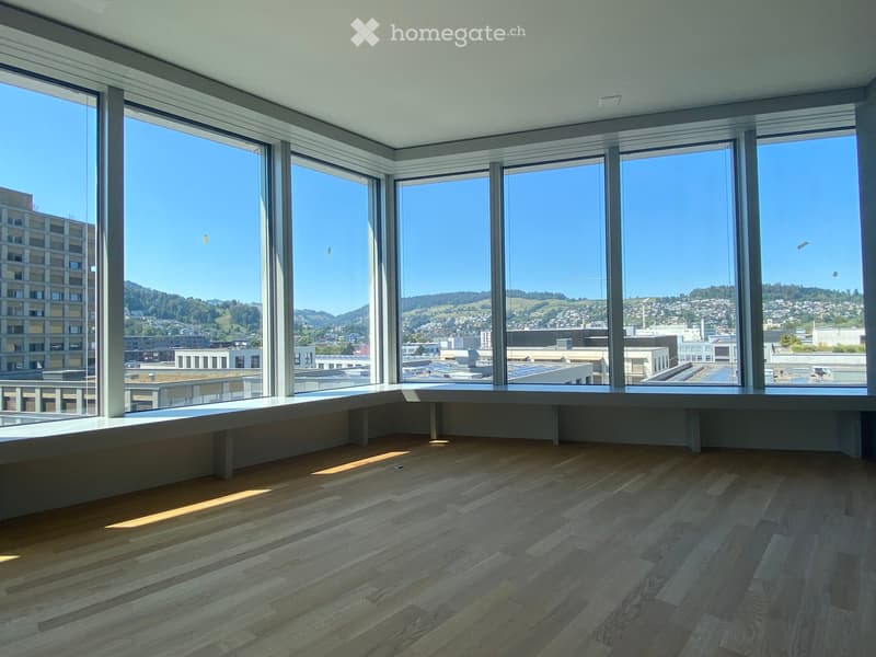 Empty room with large windows offering a cityscape view, wooden flooring, and a built-in bench along the windows.