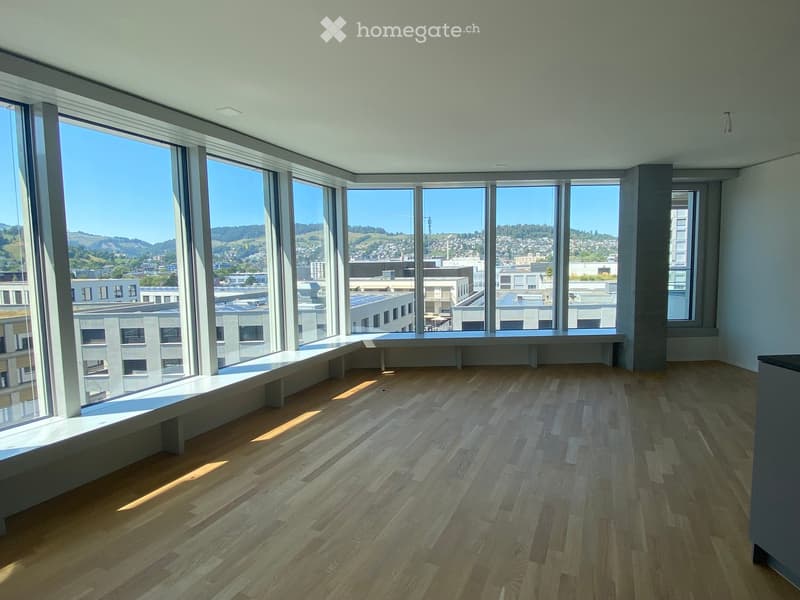 Empty room with wooden flooring and large windows offering a cityscape view. A long bench is positioned along the window wall.