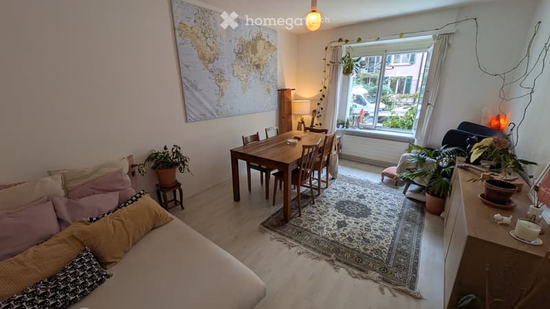 A cozy living and dining area with a sofa, dining table, chairs, potted plants, a world map, and a guitar. The space is well-lit with natural light from a large window and has a warm, inviting atmosphere.