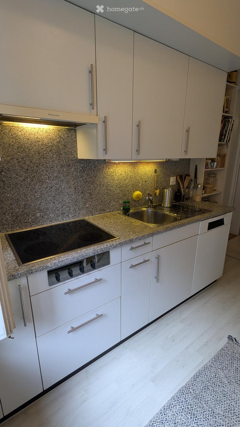 Modern kitchen with white cabinets, granite countertops, and stainless steel appliances including a stove, sink, and oven. The floor is light-colored wood, and the backsplash is granite.