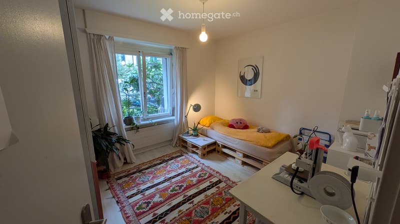 Cozy bedroom with a bed, nightstand, small table, chair, fan, lamp, plant, and picture frame on the wall.