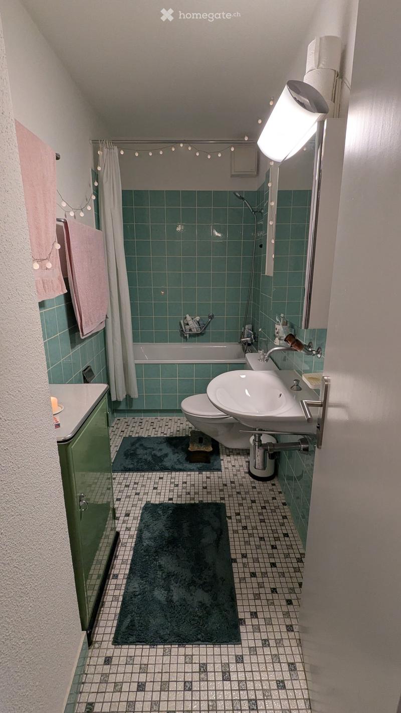 Bathroom with green tiled walls, bathtub, toilet, sink, and two rugs on the floor.