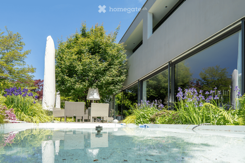 Modern outdoor area with a swimming pool, lounge chairs, umbrella, and lush garden with flowers and trees.