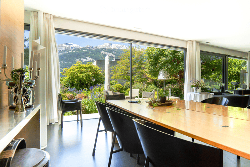 Bright room with large windows offering a scenic mountain view, furnished with a long wooden table, chairs, a decorative candle holder, and a vase on a side table.