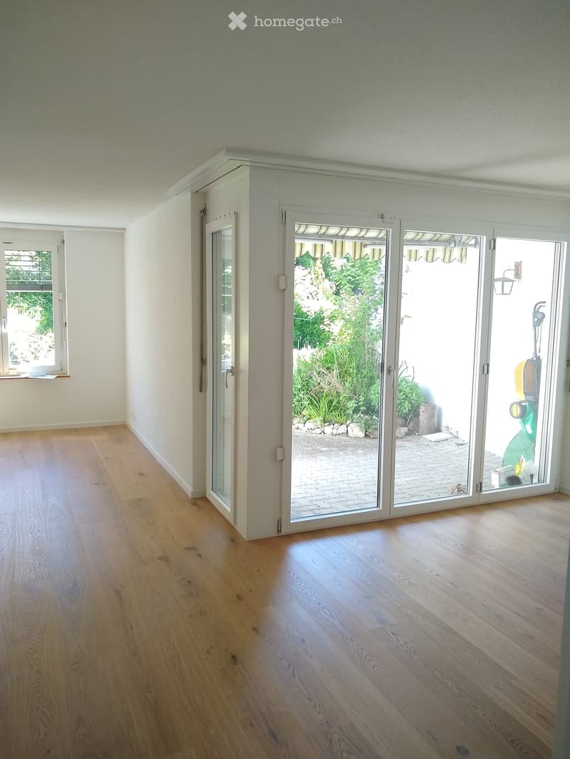 Empty room with light wooden flooring, white walls, large sliding glass doors leading to a garden, and a window on the left side.
