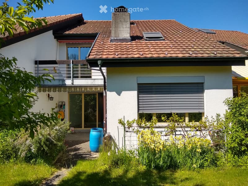 Two-story house with a tiled roof, white exterior, balcony, and garden area with plants and a blue barrel.